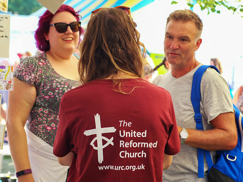 A volunteer in a United Reformed Church T-shirt chatting with visitors at an outdoor event