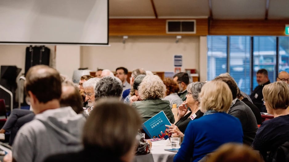 Delegates seated at round tables during a Leading your Church into Growth conference session, one holding the Leading Your Church into Growth handbook